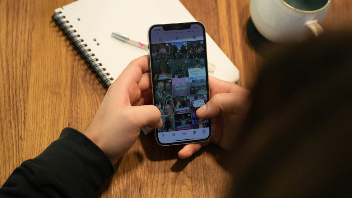 Eine Person, die auf einem Smartphone eine Social-Media-Galerie mit Bildern und Beiträgen durchscrollt, mit einem Notizblock und einer Tasse im Hintergrund, auf einem Holztisch liegend.
